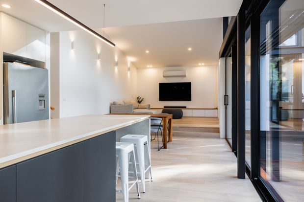 Bright open-plan contemporary interior combining a minimalist kitchen with island seating, dining area, and living room. The space features dark kitchen cabinetry, a pale benchtop, light timber flooring, white walls, recessed and linear lighting, and extensive black-framed glazing along one side.