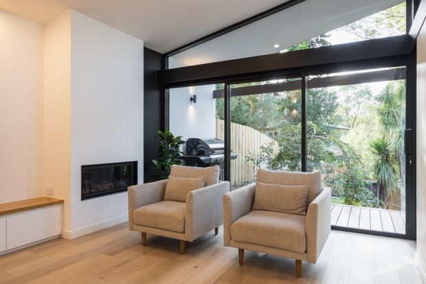 A bright contemporary living room with two upholstered armchairs arranged beside a wall-mounted linear fireplace. Full-height black-framed glazing opens onto a timber deck and leafy garden, with a high clerestory window bringing in additional natural light. The room features pale timber flooring, white walls, dark window framing, and a simple built-in cabinet.