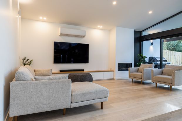 Bright contemporary living room with a large light-grey sectional sofa, two matching armchairs, pale timber flooring, white walls and recessed ceiling lighting. A wall-mounted television, low built-in media cabinetry, split-system air conditioner and compact fireplace are visible. Full-height black-framed glazing opens toward a fenced outdoor area, adding natural light to the room.