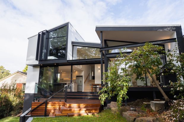 Exterior view of a contemporary multi-level residence with crisp white and dark charcoal cladding, extensive black-framed glazing, projecting box forms, and a broad flat roof. Timber steps lead to a covered entry and raised outdoor deck, with established garden planting in the foreground.