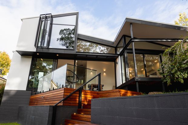 Contemporary multi-level residence viewed from the street, featuring bold geometric rooflines, extensive floor-to-ceiling glazing, white rendered walls, black-framed windows, a warm timber deck and stair, and dark masonry retaining walls. The elevated entry terrace and open-plan interior are visible through the glazing.