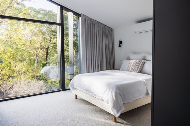 Bright contemporary bedroom with a low-profile upholstered bed, white bedding, neutral striped accent cushions, full-height black-framed glazing, and grey curtains. The room overlooks leafy natural surroundings and has a soft light-grey carpet, white walls, a wall-mounted air conditioner, and a small black wall light.