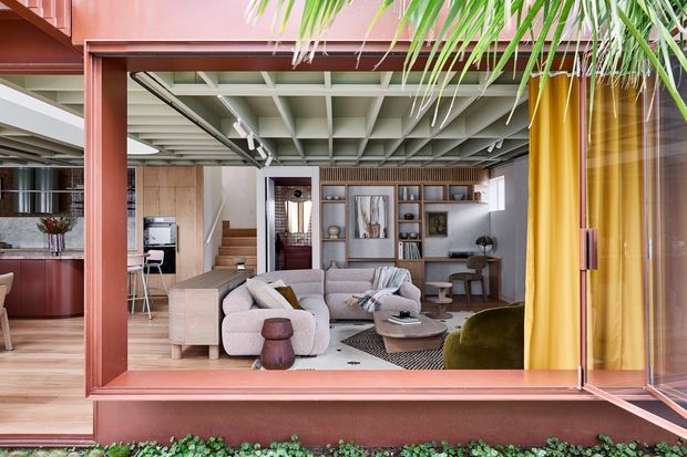 A stylish open-plan living area viewed through a large framed opening, with a pale sectional sofa, sculptural coffee table, built-in shelving, and a compact kitchen visible to the left. The room features exposed white coffered structural ceiling beams, warm timber cabinetry and flooring, and deep terracotta-red framing. Large glazed openings and tropical foliage create a strong indoor-outdoor connection. The look is refined and contemporary with mid-century influences.