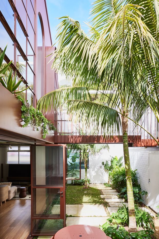A bright, tropical courtyard viewed from beside an open glazed door. The space features a narrow landscaped garden with palms and lush planting, pale stepping-stone paving, white masonry walls, and a red-brown metal-framed balcony or bridge overhead. Tall floor-to-ceiling glazing and matching dark reddish frames define the contemporary building, while a glimpse of an interior living area is visible at lower left. A round pink outdoor table occupies the foreground.