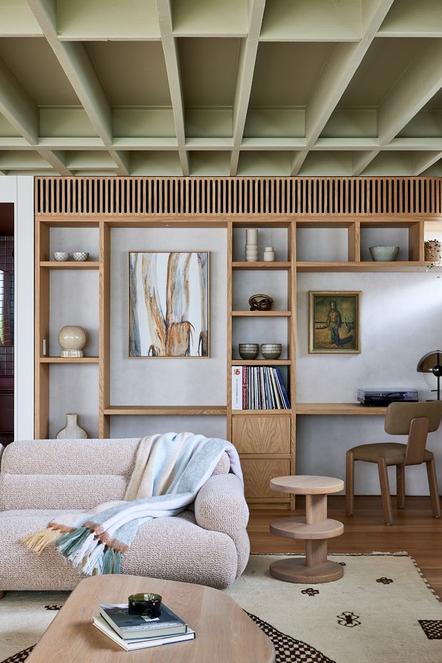 A refined living room with a large built-in timber shelving and display wall, integrated desk, open shelving, artwork and decorative objects. A textured cream sofa with a draped throw faces a rounded timber coffee table, while a smaller pedestal side table sits beside it. The ceiling features exposed painted beams arranged in a deep coffered grid. The palette is warm and neutral, with light timber, cream upholstery, pale walls and carefully styled furnishings.