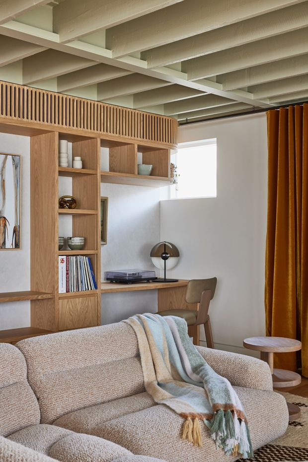 A warm, thoughtfully designed living room with a built-in timber shelving and desk unit, upholstered sofa, mid-century style desk chair, sculptural table lamp, record player, and rich mustard curtains. The exposed painted ceiling beams add architectural character, while a slatted timber upper screen creates visual separation and storage interest.