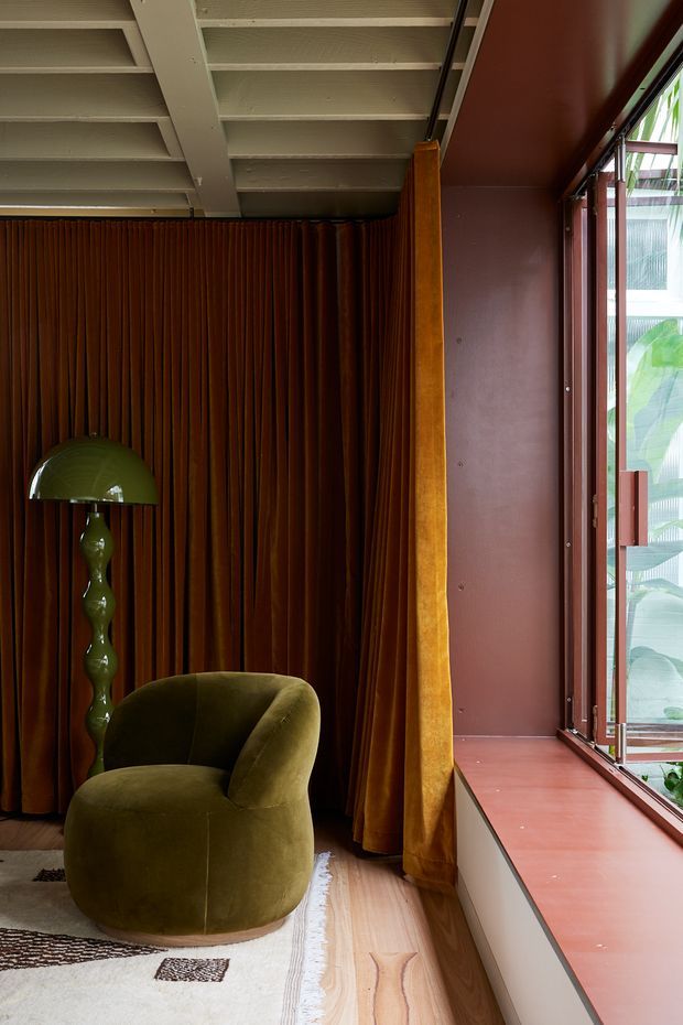 A richly styled interior corner with a rounded olive-green lounge chair and matching sculptural table lamp set against full-height dark rust-brown pleated curtains. The room has pale timber flooring, a patterned light rug, a deep reddish-brown window wall and sill, and an open glazed window overlooking leafy greenery. Exposed painted ceiling joists add architectural character, while the palette and plush forms create a warm, intimate atmosphere.