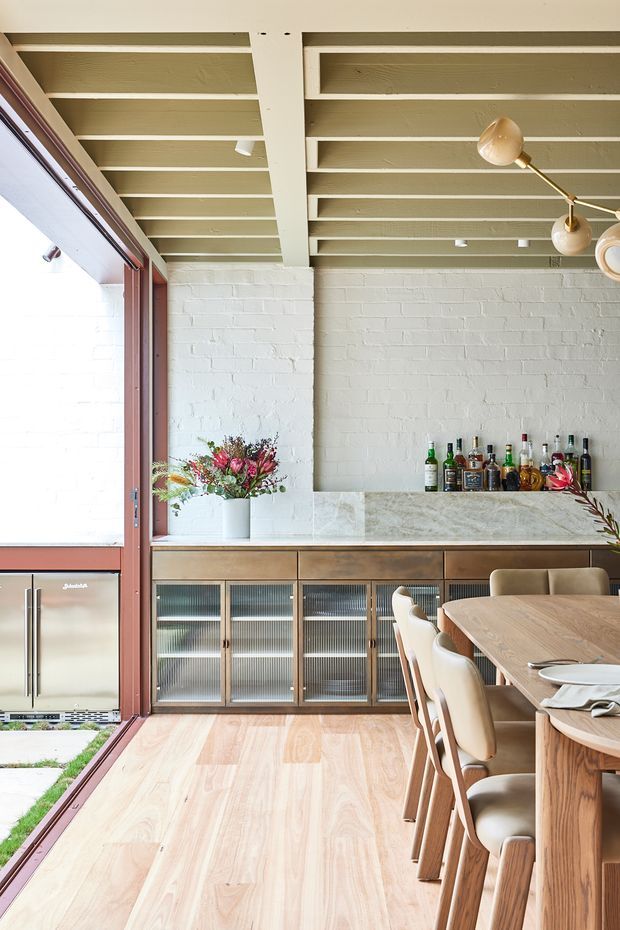 Bright open-plan dining and entertaining area with a built-in drinks bar along a white-painted brick wall. The space features a timber dining table and upholstered dining chairs, a long stone-look benchtop, timber cabinetry with ribbed glass fronts, pale timber flooring, exposed painted ceiling beams, and large burgundy-framed glazed doors opening to the outside. A compact beverage refrigerator and bottles are visible, along with a sculptural pendant light.