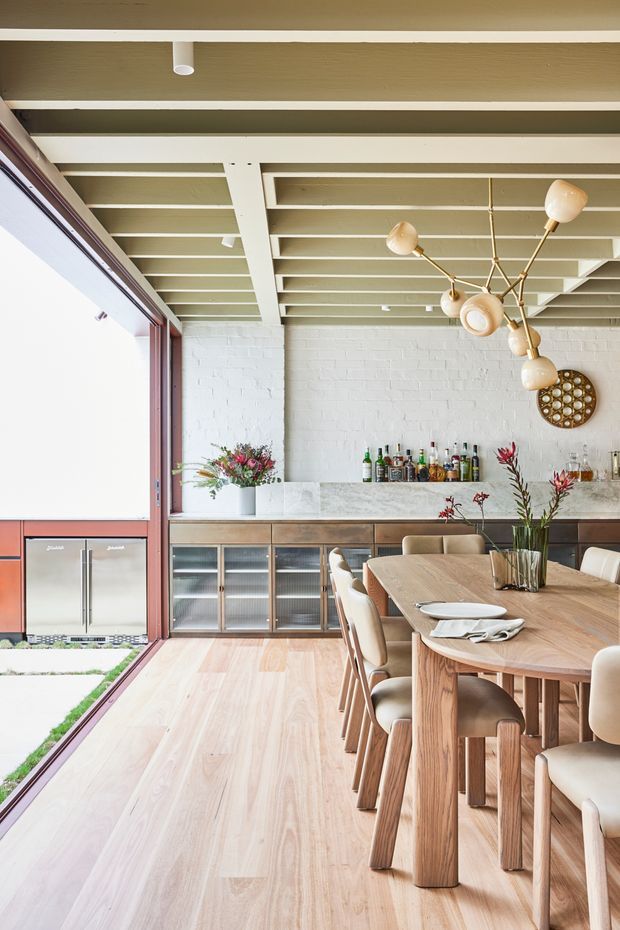 A bright open-plan dining and entertaining area with a long light timber dining table, upholstered dining chairs, and a built-in bar cabinet along the rear wall. The space features white-painted brick walls, a pale stone benchtop, timber flooring, extensive glazed openings to the left, exposed painted ceiling beams, and a sculptural branching pendant light. Bottles and glassware are arranged on the built-in bar, while floral arrangements add colour to the room.
