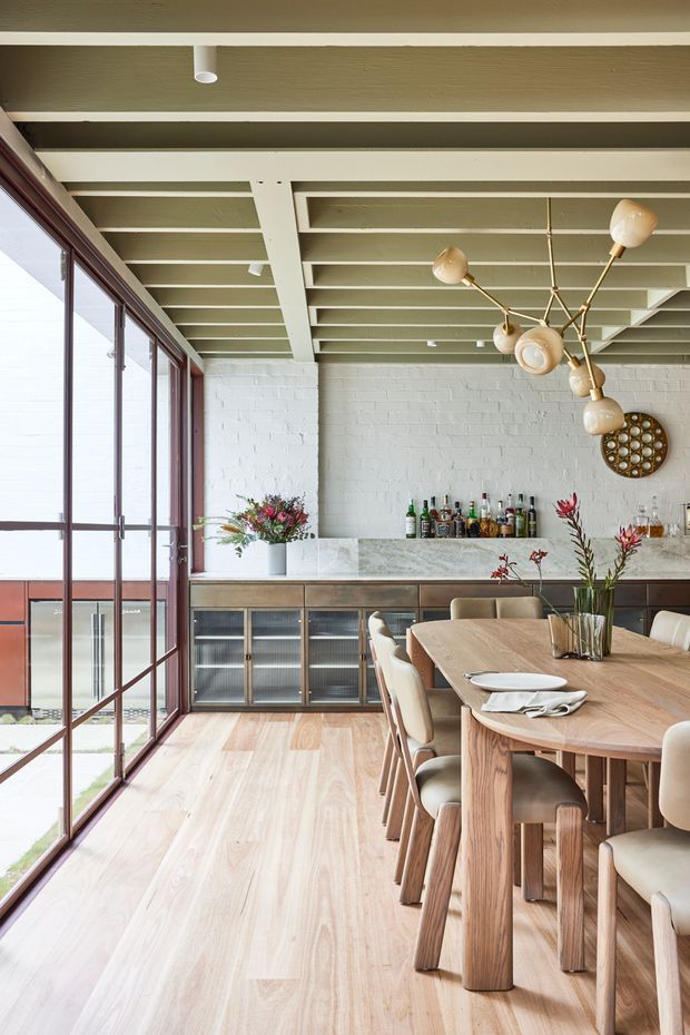 A bright, refined dining room with a long timber dining table and upholstered dining chairs beside full-height glazed doors. The room features a painted white brick wall, built-in dark timber and glass cabinetry, a pale stone benchtop, pale timber flooring, exposed painted ceiling beams, and a sculptural brass-and-glass chandelier. A drinks and bar area is arranged along the rear wall, with indoor plants and flowers adding colour.