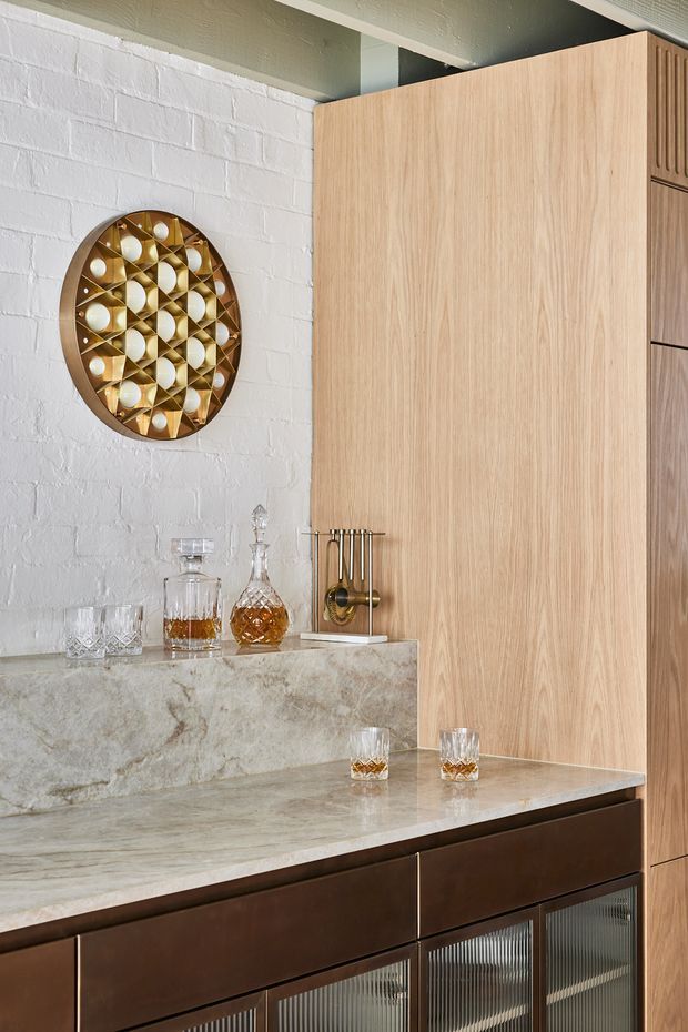 Close-up interior view of a refined home bar with dark timber cabinetry, a pale veined stone benchtop, light timber wall-height joinery, and a white painted brick wall. Glassware, decanters, spirits and a brass-toned bar tool holder sit on the counter, with decorative wall art above.