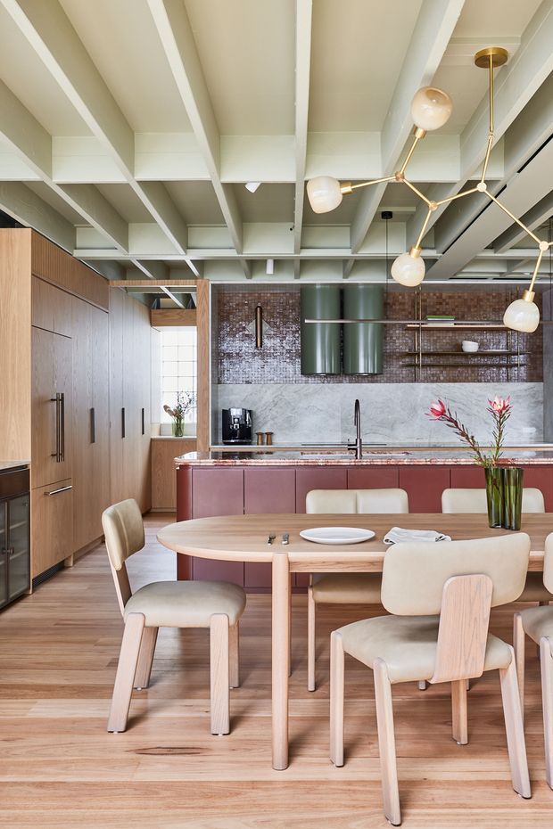 A warm, contemporary open-plan kitchen and dining area. The space features extensive mid-tone timber cabinetry and flooring, a burgundy island with a pale stone benchtop, textured dark brown tile backsplash, open shelving, and a green metallic rangehood. A round timber dining table with upholstered cream chairs occupies the foreground. The ceiling has exposed white-painted beams and joists, complemented by a sculptural brass-and-globe pendant light.