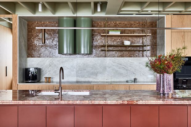 A contemporary kitchen photographed straight on, featuring a large marble island with an integrated sink and tall dark tap, muted red-brown cabinetry, light timber wall cabinetry, a textured brown brick-tile backsplash, open timber shelving, and a prominent green cylindrical rangehood. The ceiling has exposed painted structural beams and recessed services, while dark stone or concrete-look side walls frame the cooking area. A coffee machine, oven, decorative vase, and kitchenware are visible.
