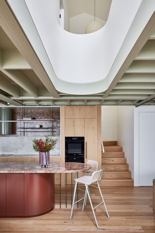 A refined open-plan kitchen and dining area with a curved reddish-brown island, patterned stone benchtop, timber cabinetry, integrated black appliances, open shelving, and a timber staircase. The space features an exposed structural ceiling grid with a large sculptural white oval ceiling opening and pendant light, creating a strong architectural focal point. Warm timber flooring and cabinetry contrast with pale walls and soft furnishings.