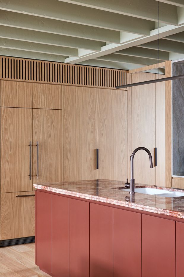 A refined contemporary kitchen centred on a large island. The island has deep terracotta-red flat-panel cabinetry, a heavily veined pink-toned natural stone benchtop, an undermount white sink, and a dark arched gooseneck tap. Full-height light timber cabinetry lines the rear wall, with integrated appliances and long dark handles. Above, a slatted timber ventilation or screening detail sits beneath exposed white painted ceiling beams. The palette is warm and earthy with strong timber grain and contrasting dark hardware.