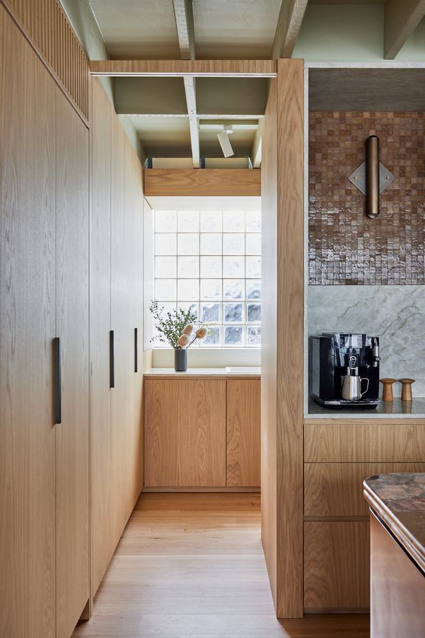 A compact, warm-toned kitchen interior viewed along a narrow galley passage. Floor-to-ceiling light timber cabinetry lines the left side, while a timber-framed coffee station with dark stone and textured brown tile surrounds occupies the right. A glass-block window brings diffused daylight into the space, with exposed structural ceiling beams and a ceiling fan visible above. The image has a refined contemporary character with strong natural wood detailing.