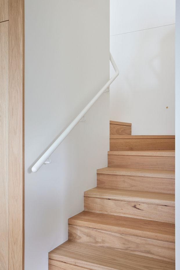 A minimalist residential stairway viewed from below, featuring broad natural timber stairs, smooth white walls, a simple white handrail, and light timber wall trim or cabinetry at the left edge. The composition is bright, clean, and understated, with no visible furnishings.