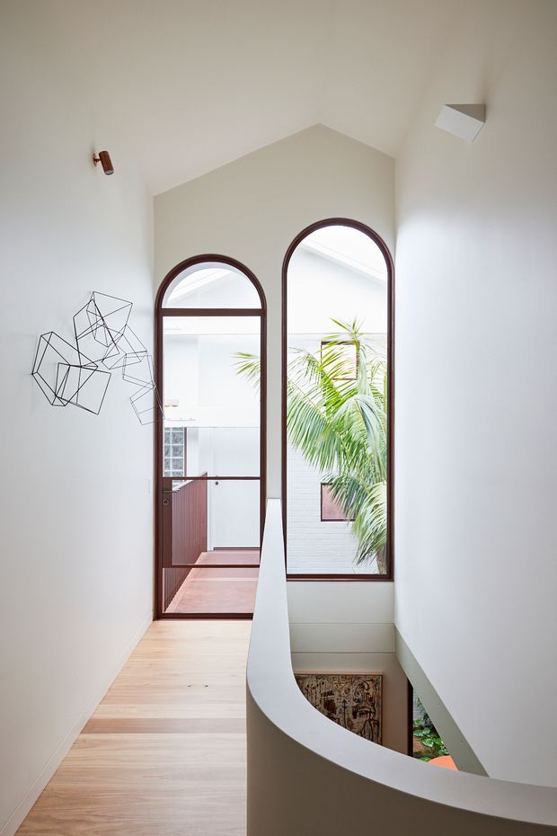 A bright, narrow upper-level landing or hallway with a dramatic curved stair balustrade and double-height ceiling. Pale timber flooring and white plaster walls are paired with tall arched timber-framed glazing and a matching arched door, overlooking an internal courtyard with tropical planting. A geometric black wire artwork decorates the wall, while a patterned feature surface is partially visible below the stair.