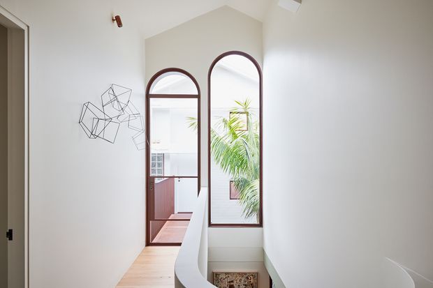 A bright, minimalist stair landing or hallway with a high vaulted ceiling, white walls, pale timber flooring, and a sculptural curved white balustrade. Two tall arched windows with dark timber frames overlook a leafy courtyard, while geometric wire wall art provides a subtle decorative feature.