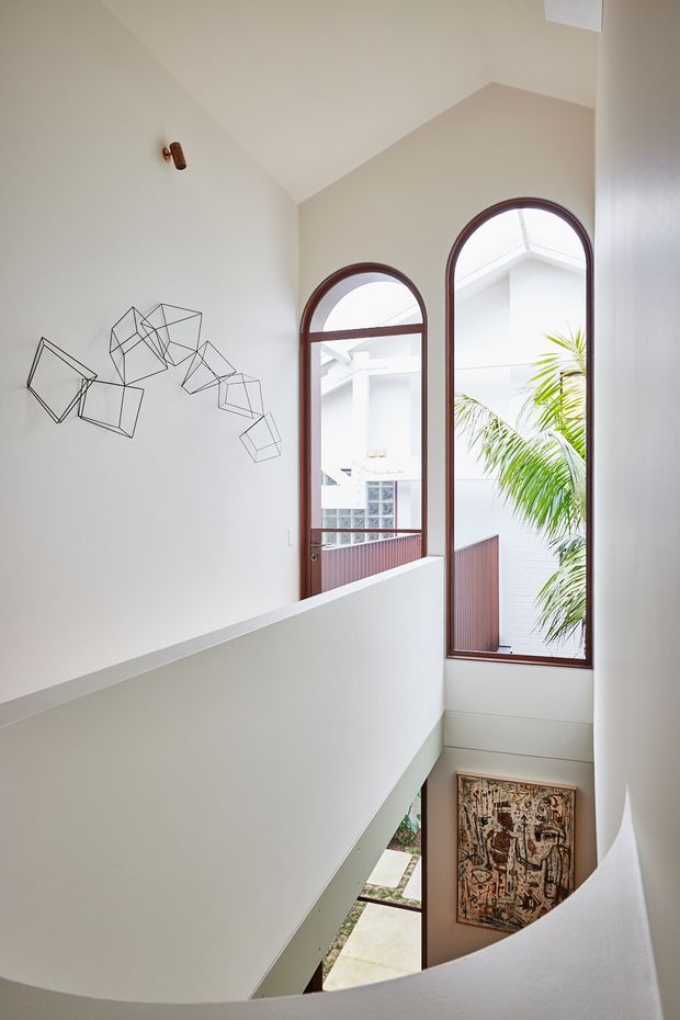 Bright architectural interior view of a stairway or landing with a curved white balustrade, double-height void, and tall arched timber-framed windows. The windows overlook an internal courtyard or adjacent building, while a minimalist geometric wire wall sculpture and framed artwork add restrained decoration. The space has strong natural light, white plaster surfaces, and a refined contemporary character.