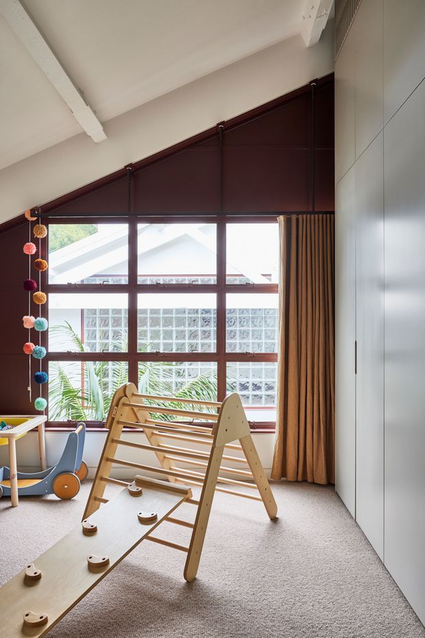 A bright children's playroom with a pitched ceiling and exposed white structural beam. The room features a large dark timber-framed window wall with glass-block and leafy views beyond, full-height warm brown curtains, pale carpet, and white built-in cabinetry. Wooden climbing and play equipment occupies the foreground, with a small colourful play table and hanging pom-pom decoration at the left.