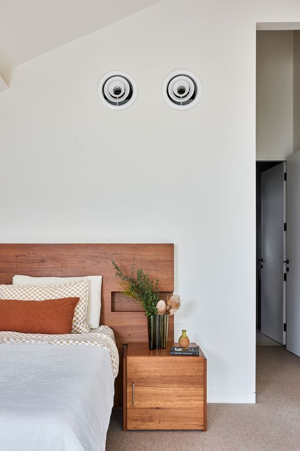 A contemporary bedroom with a warm timber feature headboard and matching built-in bedside table. The bed is dressed in white and rust-orange textiles with patterned cushions, while two circular wall-mounted fixtures provide a distinctive architectural detail above the bed. A doorway opens to a darker adjacent space, and beige carpet covers the floor.