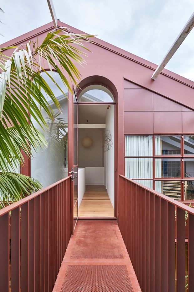 A narrow elevated pedestrian bridge leads to an open arched doorway in a striking pink-red metal-clad building. The bridge has matching vertical metal balustrades and perforated metal decking. Through the doorway, a compact interior hall with pale walls, timber flooring, and a spherical pendant light is visible. Large framed glazing sits to the right, while tropical palm foliage partially screens the structure. The image has a vivid contemporary architectural character with strong geometric lines and an indoor-outdoor connection.