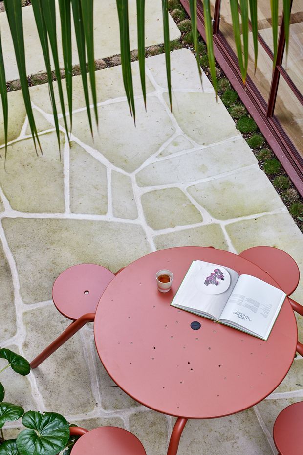 A small outdoor patio or terrace paved with irregular pale stone slabs and light mortar joints. A rounded coral-red metal table with matching stools occupies the foreground, holding an open book and a small cup. Tall narrow green leaves partially frame the view, while red-framed glazing or doors appear along the right edge. The setting feels intimate, colourful, and garden-like.