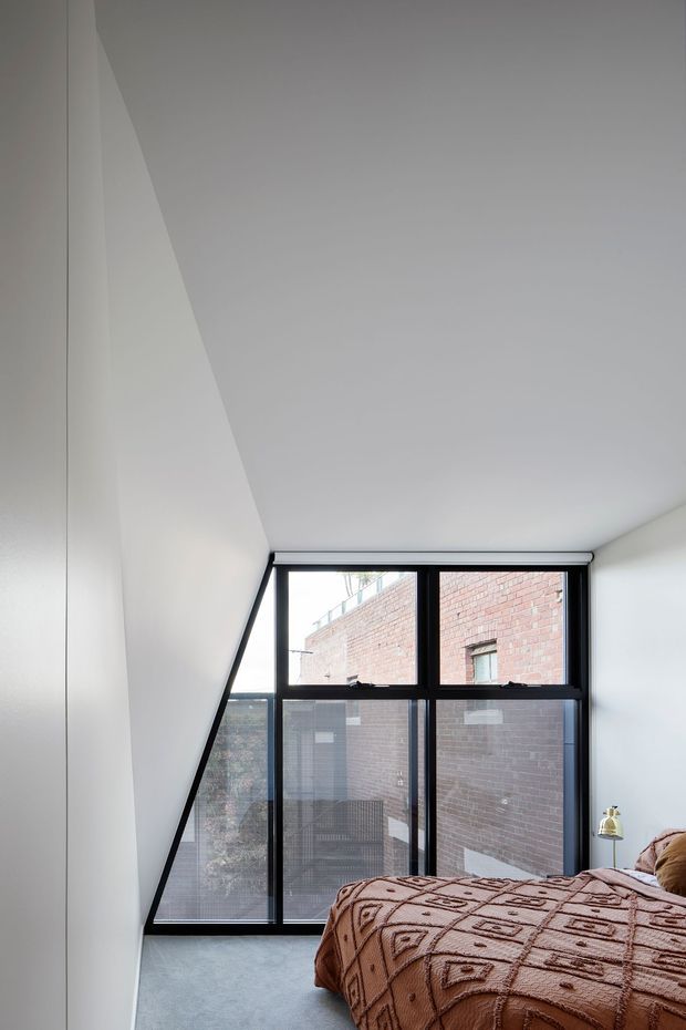 A minimalist bedroom with a low, sloping white ceiling and a dramatic full-height black-framed window wall. The glazing looks onto a close brick building, while a bed with a warm brown patterned cover occupies the foreground. The room has a restrained neutral palette, abundant natural light, and strong geometric architectural lines.