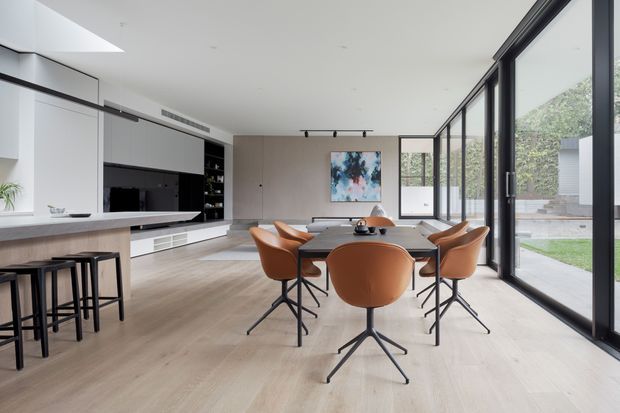 A bright, open-plan contemporary kitchen and dining area with extensive floor-to-ceiling black-framed glazing along one side. The kitchen features white and dark built-in cabinetry, a pale stone-look island bench with black bar stools, and integrated appliances. A timber dining table with sculptural tan leather chairs occupies the centre, with light timber flooring, a large abstract artwork, and views to a landscaped courtyard and pool area beyond the glazing.