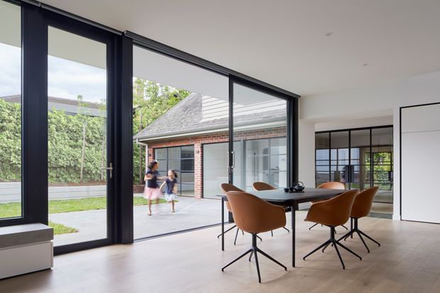 A bright, contemporary dining area with a rectangular dark dining table and sculptural tan leather chairs, opening through expansive black-framed glazing to a paved courtyard. The space has pale timber flooring, white walls and ceiling, large sliding glass doors, and a black-framed glazed internal partition leading to another room. A brick outbuilding and landscaped courtyard are visible beyond the glazing.