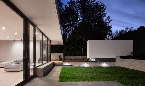 A dusk view of a contemporary house opening onto a landscaped courtyard. Full-height dark-framed glazing and sliding doors reveal a softly lit minimalist interior, while the exterior features a paved terrace, raised lawn, low rendered walls, timber screening, and integrated landscape lighting.