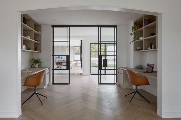 A bright, symmetrical home office or study viewed through a wide opening, with two built-in desk and shelving workstations facing each other. The room features white walls and cabinetry, warm timber herringbone flooring, black-framed glazed internal doors, and contemporary tan leather swivel chairs. A glazed partition leads to an additional furnished area beyond, while plants and books add restrained decoration.