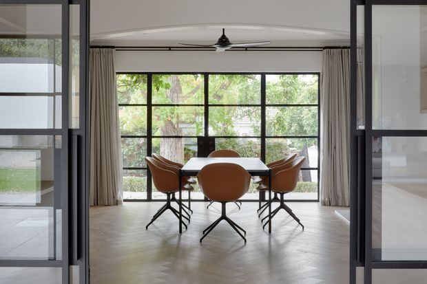A refined dining area viewed through open black-framed glazed doors. A rectangular dark dining table is surrounded by sculptural caramel-brown chairs, set against full-height black-framed windows with pale curtains and leafy garden views. The room has a pale herringbone timber floor, white walls and ceiling, slim dark ceiling detailing, and a central black ceiling fan. The overall composition is calm, contemporary, and strongly connected to the garden.