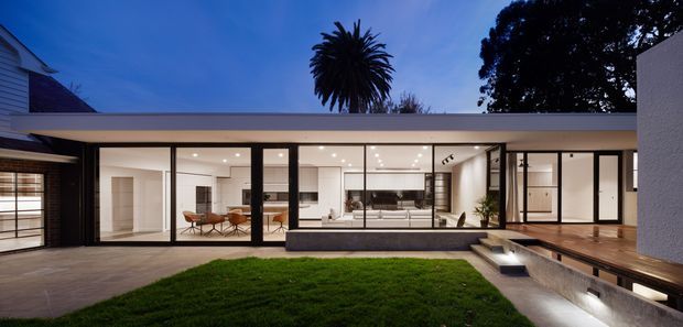 Twilight view of a low, flat-roof contemporary residence enclosing a landscaped courtyard. Extensive floor-to-ceiling glazing with dark slender frames reveals a bright open-plan interior, while a timber deck, linear water feature or pool edge, and lawn occupy the foreground. Mature trees and neighboring residential forms frame the composition.