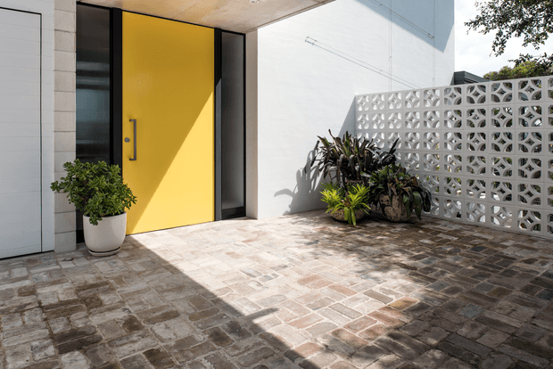Contemporary residential entry courtyard featuring a vivid yellow front door set within dark-framed glazing, pale rendered walls, and a decorative white breeze-block screen. The paved forecourt uses varied earthy-toned rectangular pavers, with potted and planted greenery adding a natural accent. Strong sunlight creates pronounced shadows across the entry.