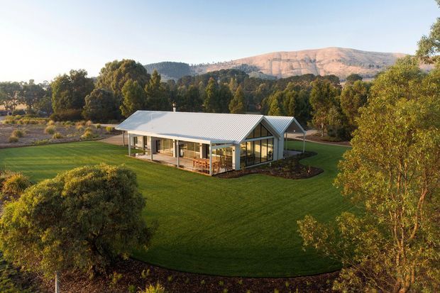 Aerial view of a single-storey contemporary pavilion-style building set within expansive manicured lawns and landscaped gardens. The building has a light grey corrugated pitched roof, extensive glazing, pale walls, and a covered outdoor terrace. Mature trees and a rugged mountain ridge form the background.