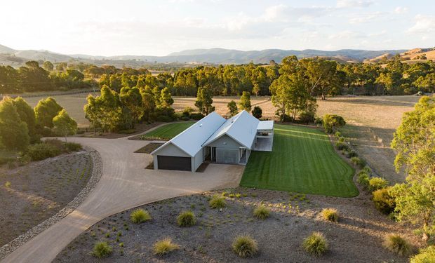 Aerial view of a contemporary single-storey rural home with multiple pitched roof forms, dark garage doors, and a pale exterior. The house sits within a landscaped property featuring a curved gravel driveway, expansive manicured lawn, native-style planting, surrounding farmland, and distant rolling hills.