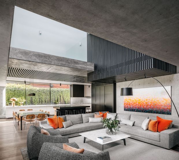 A sophisticated open-plan living and dining area with a contemporary concrete structure, exposed concrete ceiling, expansive glazing, and a dramatic black slatted mezzanine or ceiling feature. A large grey sectional sofa, textured armchairs, orange accent cushions, a white coffee table, and abstract artwork create a refined yet warm interior. The dining area and kitchen are visible in the background, opening onto a planted courtyard.