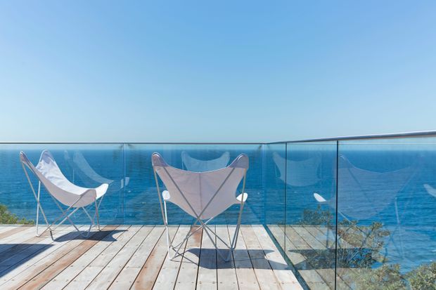 A bright coastal balcony or terrace with a weathered timber deck, transparent glass balustrade, and two folding sling chairs facing a vivid blue ocean. Low coastal vegetation is visible below the railing, with an expansive clear sky beyond.