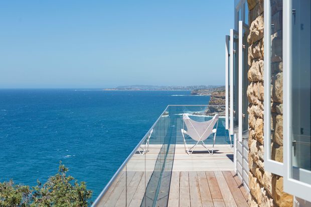 A sunlit coastal balcony or terrace projects over a vivid blue sea, with a transparent glass balustrade, timber decking, and a rugged natural-stone exterior wall with open white-framed windows. Outdoor sling chairs sit at the far end, while a distant shoreline is visible across the water.