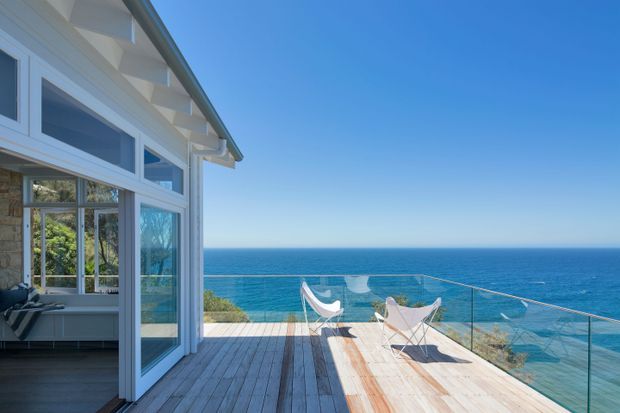 Exterior view of a coastal home opening onto a spacious timber deck overlooking the ocean. The white-framed glazed facade includes projecting clerestory-style windows and visible exposed eaves, while a frameless glass balustrade defines the deck edge. Minimal white outdoor lounge chairs occupy the sunlit terrace.