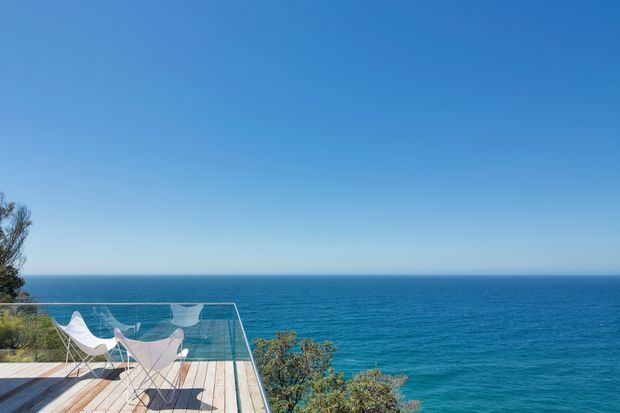 A bright coastal exterior view of a timber deck or terrace projecting toward an expansive blue ocean. The deck has a transparent glass balustrade, sculptural white outdoor lounge chairs, and sparse coastal vegetation along the edges. The image is dominated by open sea and clear sky, creating a minimalist, elevated outdoor setting.
