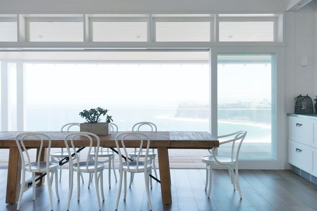 Bright dining area with a long rustic timber dining table and multiple white bentwood-style chairs, positioned in front of expansive windows and glazed doors overlooking a coastal seascape. White walls, pale flooring, simple cabinetry, and abundant natural light create an airy, relaxed interior.