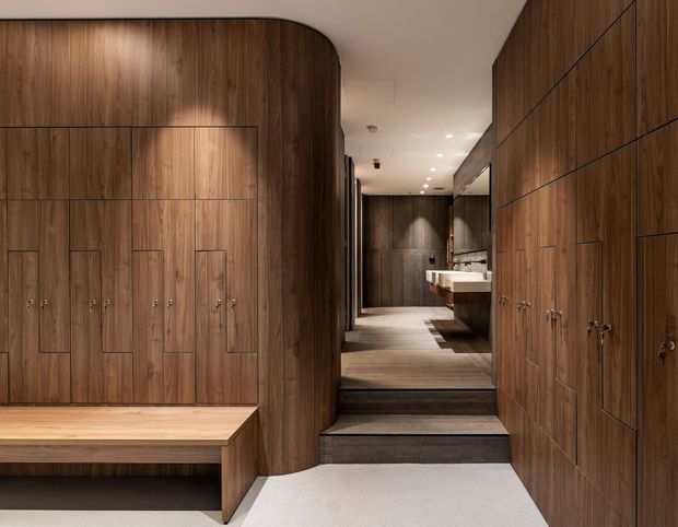 A refined, dark timber-lined interior hallway or dressing area with extensive built-in cabinetry, integrated handles, and a built-in bench. The space leads up a short set of steps toward a bathroom vanity visible at the rear. Warm recessed lighting, curved timber wall detailing, and dark stone or timber stair treads create a sophisticated, luxurious atmosphere.