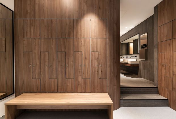 A refined walk-in wardrobe or dressing area with full-height dark timber cabinetry featuring integrated vertical doors and concealed handles. A light timber built-in bench sits below the cabinetry, while stepped flooring leads toward a vanity or bathroom area visible in the background. The space has warm, layered lighting and a restrained contemporary design.