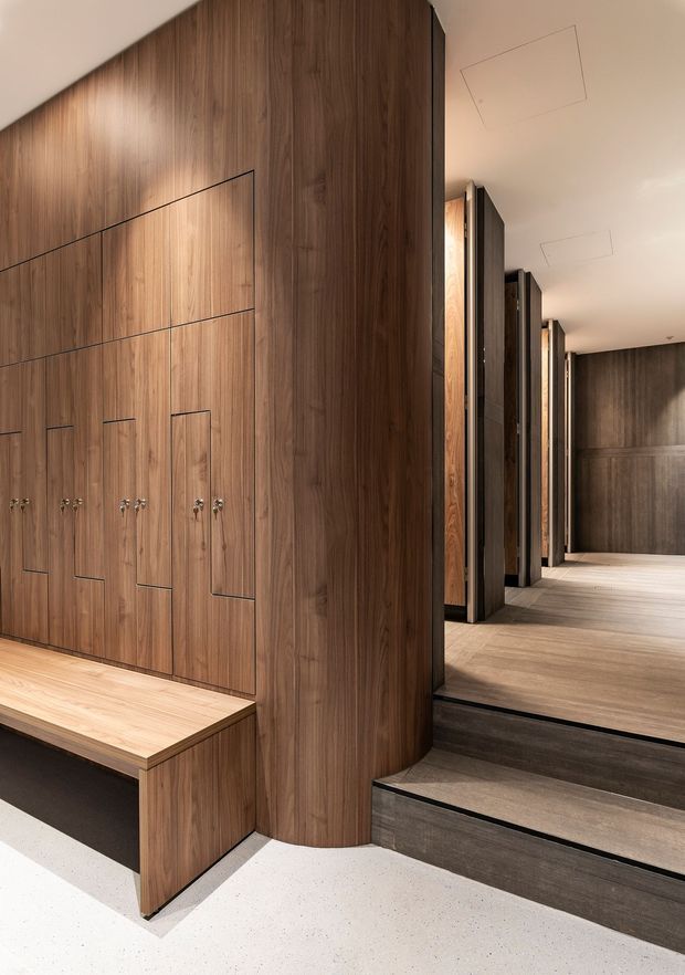 A refined contemporary interior corridor featuring extensive dark walnut-toned timber wall panelling and integrated storage with discreet vertical handles. A built-in timber bench projects from the wall, while dark stone or tiled steps lead to a raised hallway lined with repeated timber and dark-framed architectural partitions. The floor is a light terrazzo-like finish, and recessed ceiling lights provide warm, understated illumination.