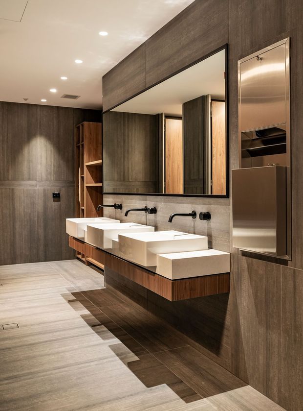 A refined contemporary bathroom with a long floating timber vanity, three white rectangular vessel basins, wall-mounted black tapware, and a large frameless mirror. Dark timber-look wall panelling and cabinetry contrast with light stone-look floor and benchtops. Recessed ceiling lights provide warm, even illumination, while a tall stainless-steel built-in storage or appliance unit is integrated into the wall.