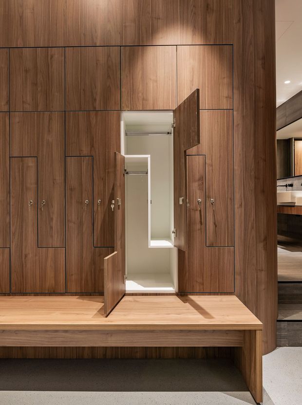 A refined built-in timber wardrobe wall with multiple flush-panel doors and integrated pulls, shown open to reveal a compact hanging and storage compartment. The cabinetry sits above a long timber bench, with warm wood flooring and dark flooring below. A glimpse of an adjacent vanity or bathroom area is visible at right. The composition is minimalist and highly tailored, with warm walnut-toned timber and concealed storage.