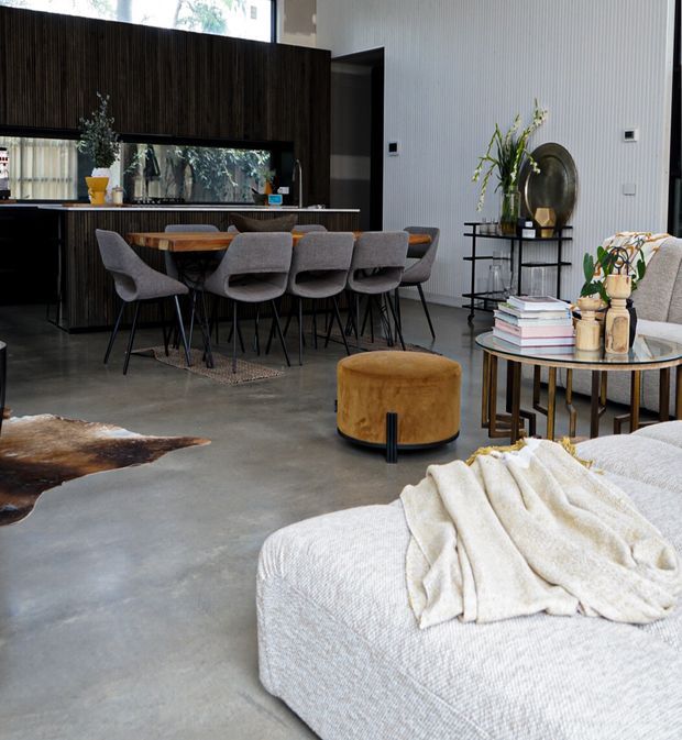 Open-plan contemporary living and dining area with a dark timber kitchen, island dining table, sculptural upholstered dining chairs, polished concrete flooring, and a light textured sectional sofa. A round ottoman, layered side tables, plants, artwork and decorative objects create a refined, warm interior.
